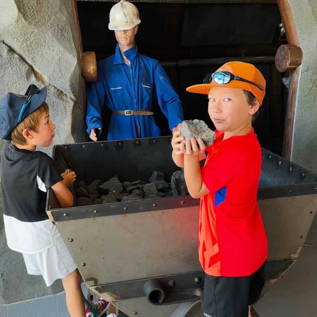 Two boys interacting with a coal cart display inside a mining-themed exhibit, with one holding a rock and a mannequin dressed as a miner behind them.