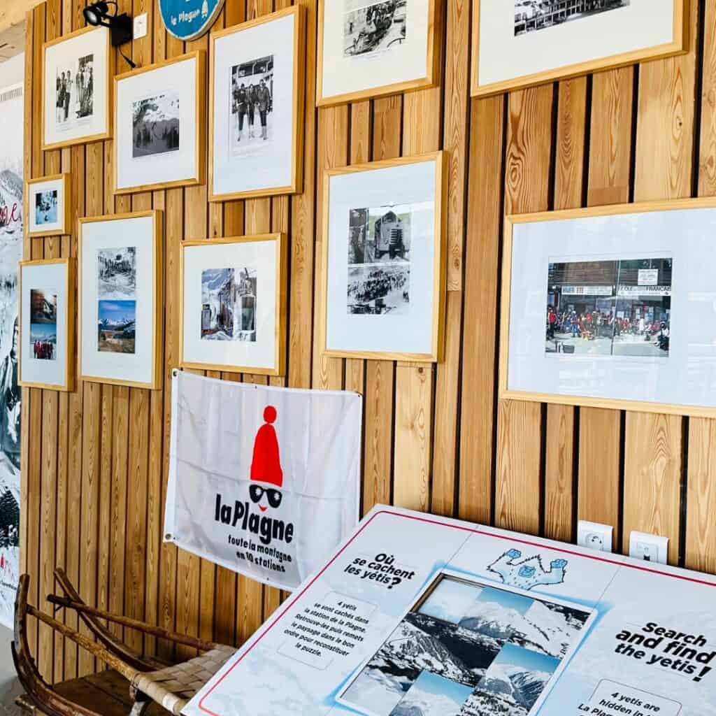 Wood-paneled museum wall featuring framed historical photos of La Plagne, a flag with the resort’s logo, and a display board inviting visitors to “Search and find the yetis.”