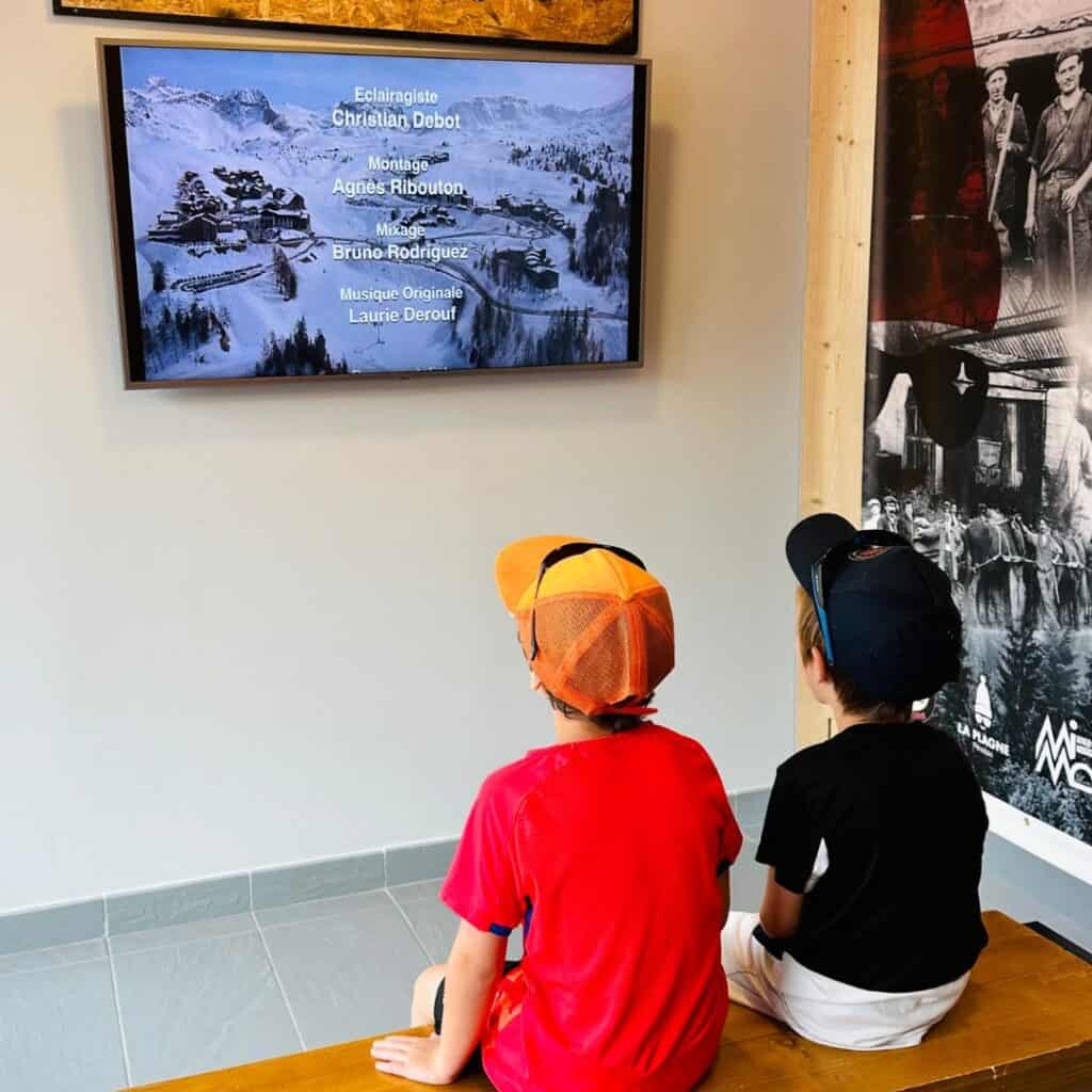 Two children seated on a bench watching a documentary or film credits on a wall-mounted screen inside a La Plagne heritage exhibit.