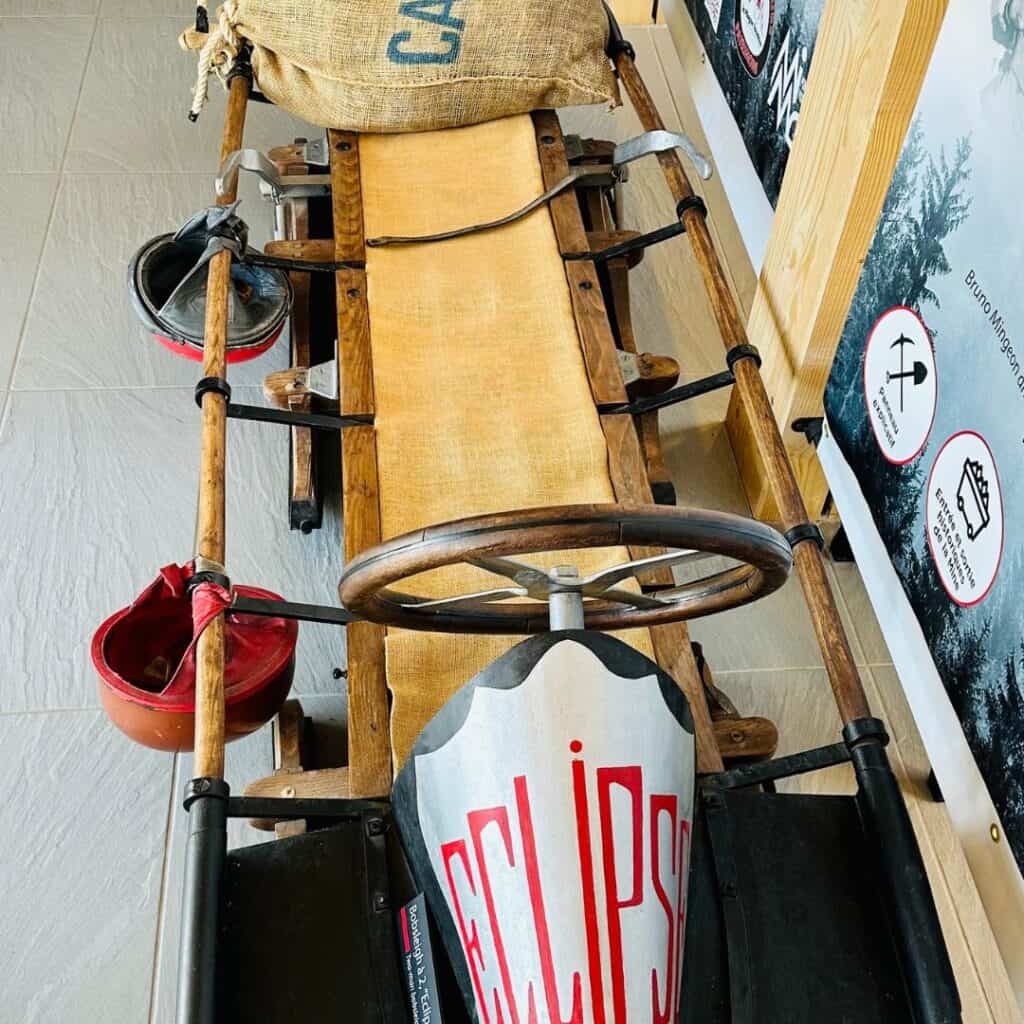 Vintage wooden bobsled labeled “Eclipse,” with a burlap sack, red helmets, and metal steering wheel, on display in a winter sports museum.