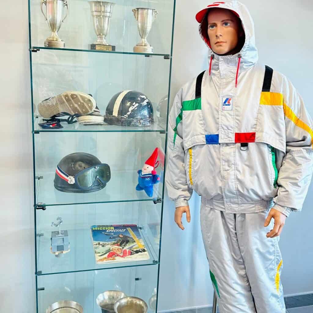 Glass display case filled with ski trophies, helmets, and memorabilia next to a mannequin dressed in retro ski gear at a winter sports exhibit.
