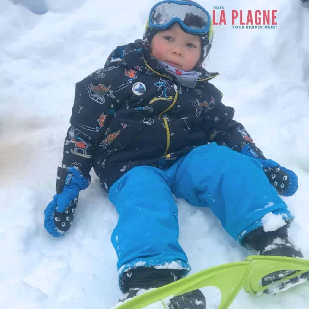 Young child bundled up in a patterned ski jacket and bright blue snow pants, sitting in the snow with a green sled in front. The child wears blue ski gloves and goggles, with rosy cheeks, under the text “That’s La Plagne – Your Insider Guide.”