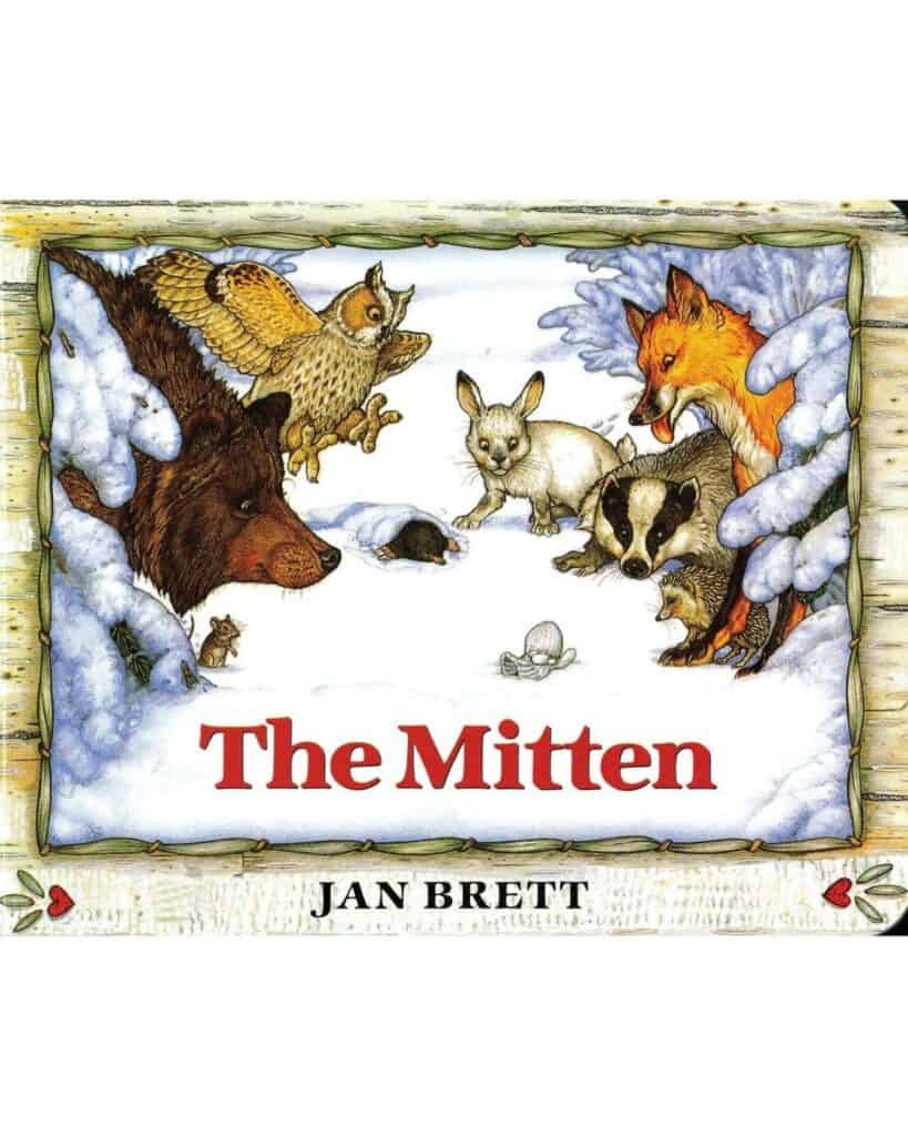 The Mitten by Jan Brett: Detailed, classic-style illustration of woodland animals—including a bear, owl, fox, rabbit, and badger—gathered curiously around a single white mitten nestled in the snow.