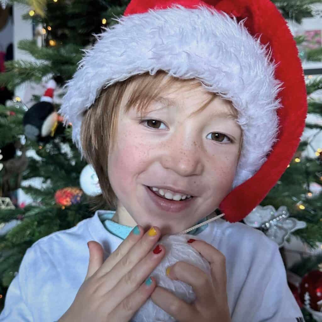 A cheerful child wearing a red and white Santa hat smiles in front of a decorated Christmas tree, holding a white ornament and showing off colorful, painted fingernails. The background features twinkling lights and holiday decorations, capturing a joyful holiday vibe.