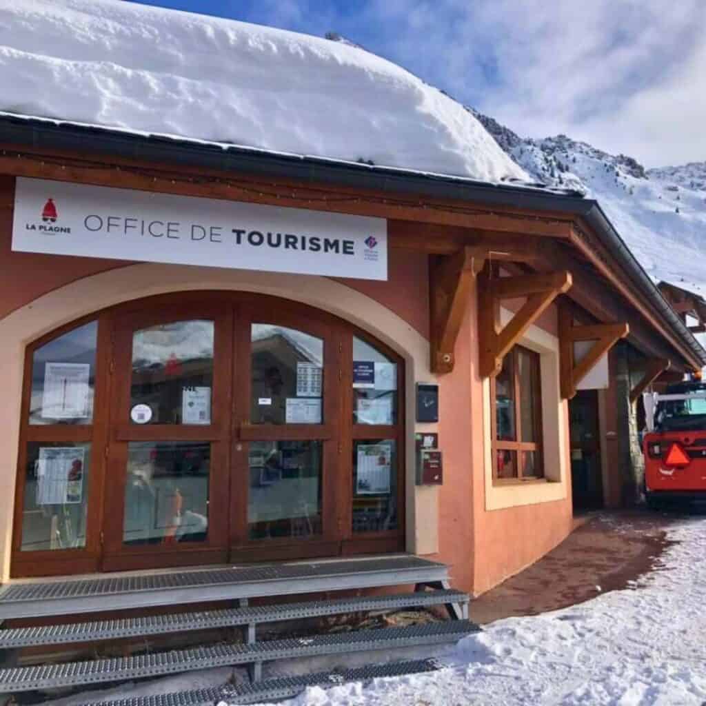 A snowy exterior view of the Office de Tourisme (Tourist Office) in La Plagne, France, with a wooden entrance, arched windows, and a sign above the door. A snow-covered roof and a red snow vehicle are visible, with alpine mountains in the background under a bright blue sky.