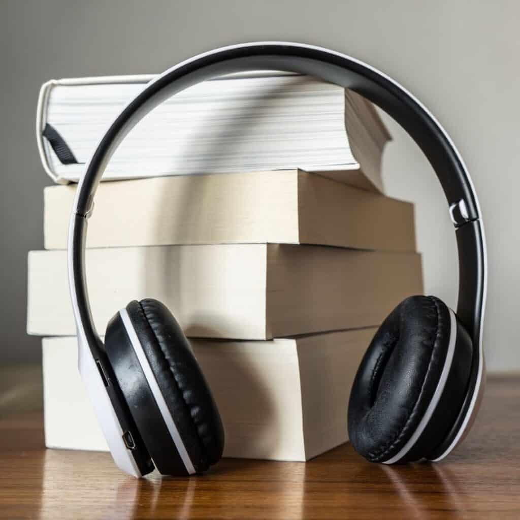 A stack of hardcover books sits on a wooden surface, with a pair of black and white over-ear headphones leaning against them, symbolizing the concept of audiobooks or learning through listening.