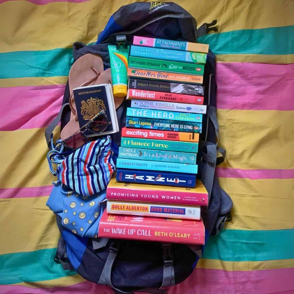A travel backpack lies open on a colorful striped bedspread, filled with a tall stack of vibrant books, a striped face mask, flip-flops, a tube of SPF 50 sunscreen, sunglasses, and a British passport. The visible book titles include The Wake-Up Call, Good Material, Promising Young Women, Hamnet, The Herd, Exciting Times, and more, suggesting a well-prepped literary getaway.