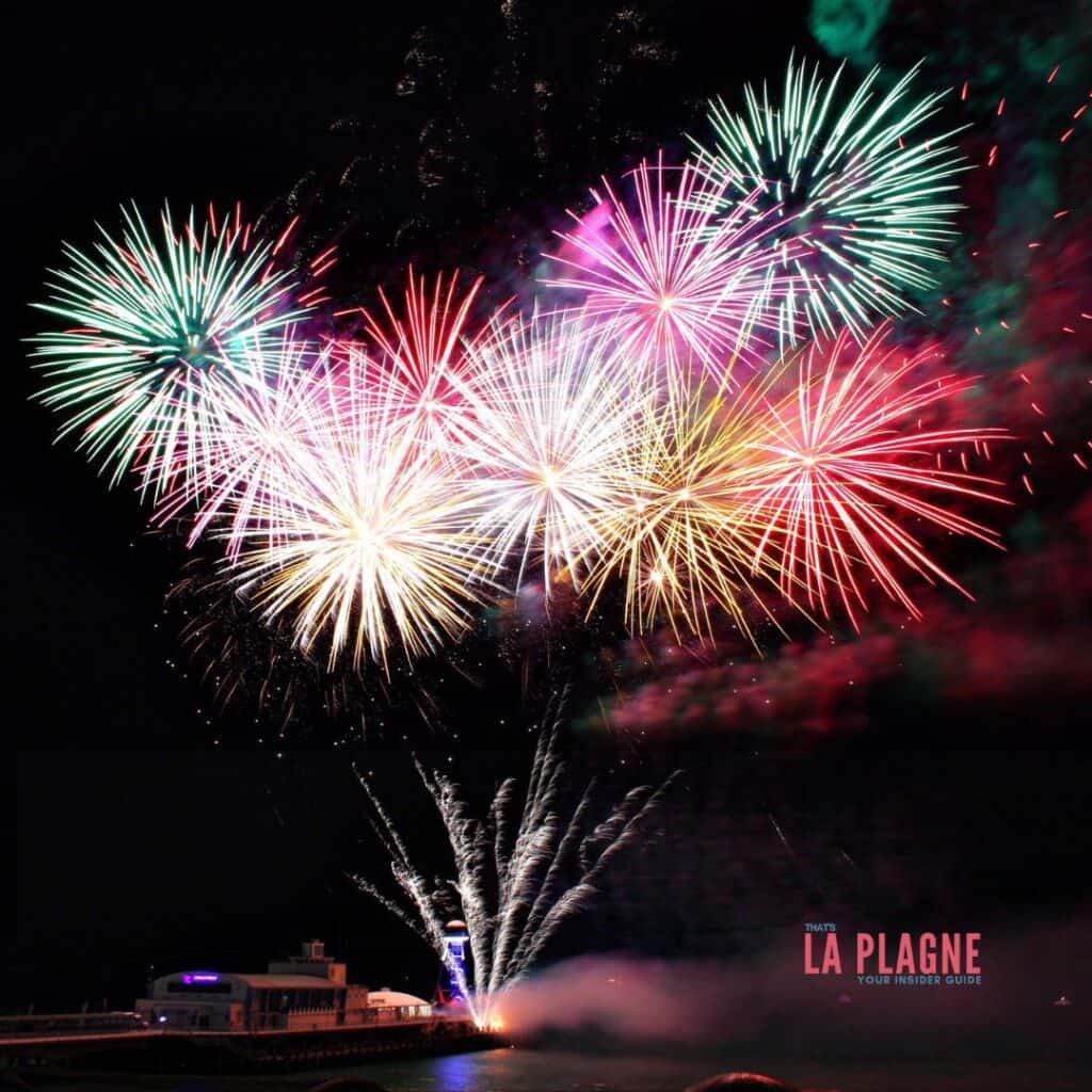 Explosive firework display in red, green, and white over a waterfront with a pier structure in view. The sky is illuminated by a dramatic spread of color with the "That's La Plagne – Your Insider Guide" logo in the corner.