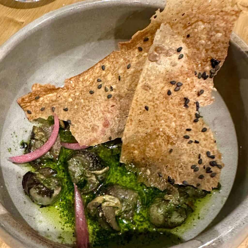 Escargot in a vibrant green herb oil, accented with pickled red onions and paired with crispy sesame flatbread. A modern twist on a French classic, served with artistic flair.