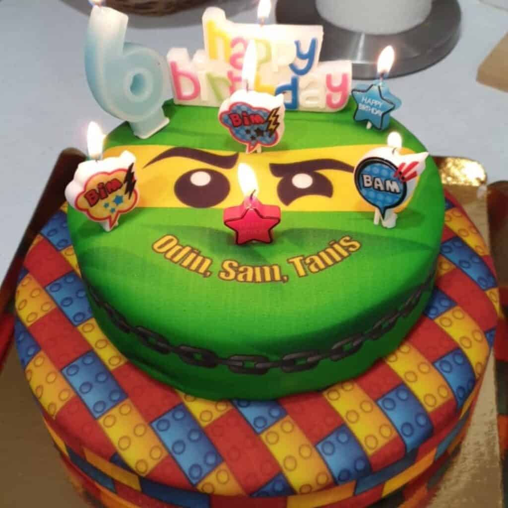 Bright and colorful two-tiered birthday cake with a Lego brick pattern on the bottom and a ninja face on top. Candles and comic-style toppers decorate the cake, which is labeled with the names “Odin, Sam, Tanis.”