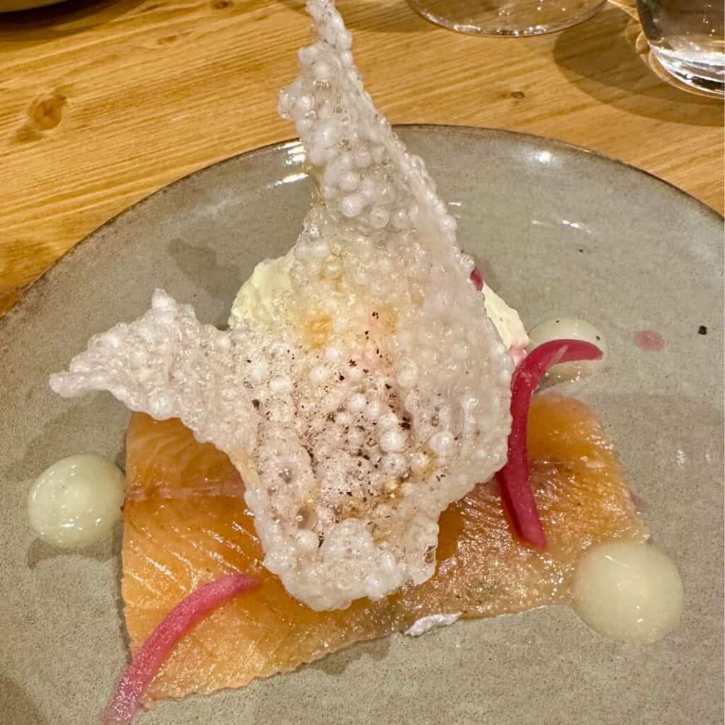 Delicate slices of cured salmon topped with pickled onions and a dramatic puffed rice cracker, plated with dots of a pale citrus or cream sauce. A textural and visual standout.
