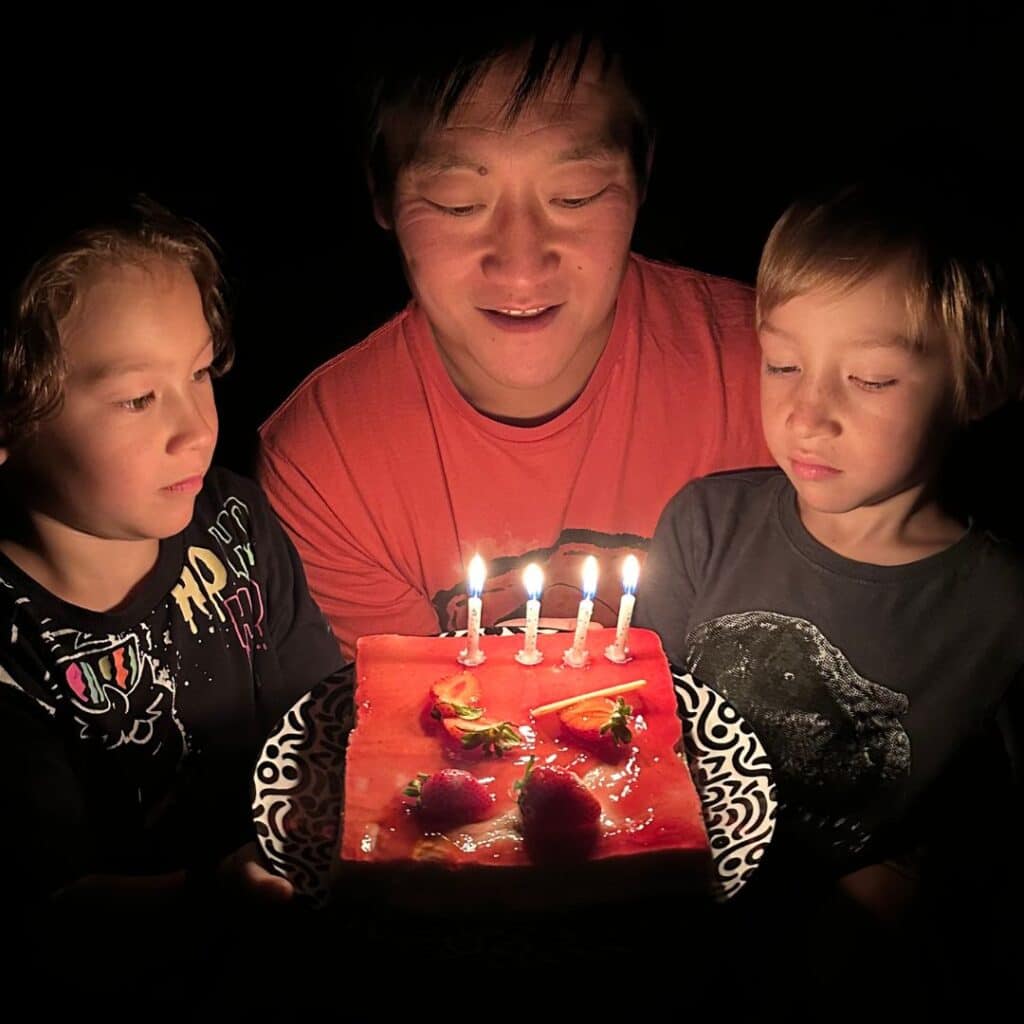Two children and an adult gather in the dark around a lit strawberry-topped birthday cake, illuminated only by the glow of five candles. The mood is sweet and intimate, capturing a quiet birthday moment.