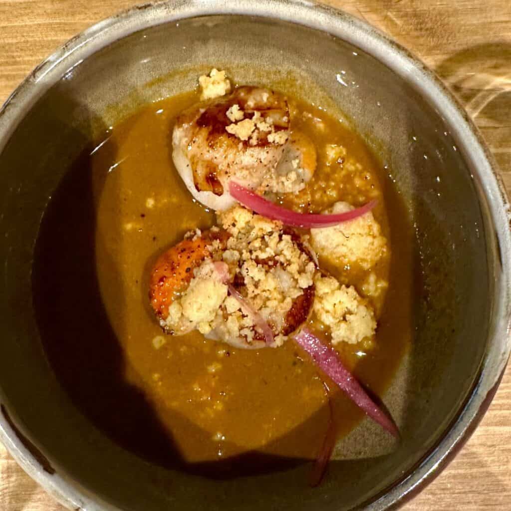 Golden-seared scallops in a velvety brown sauce, topped with crumbled biscuit or streusel and pickled onions. A cozy yet elegant dish with sweet and savory layers.