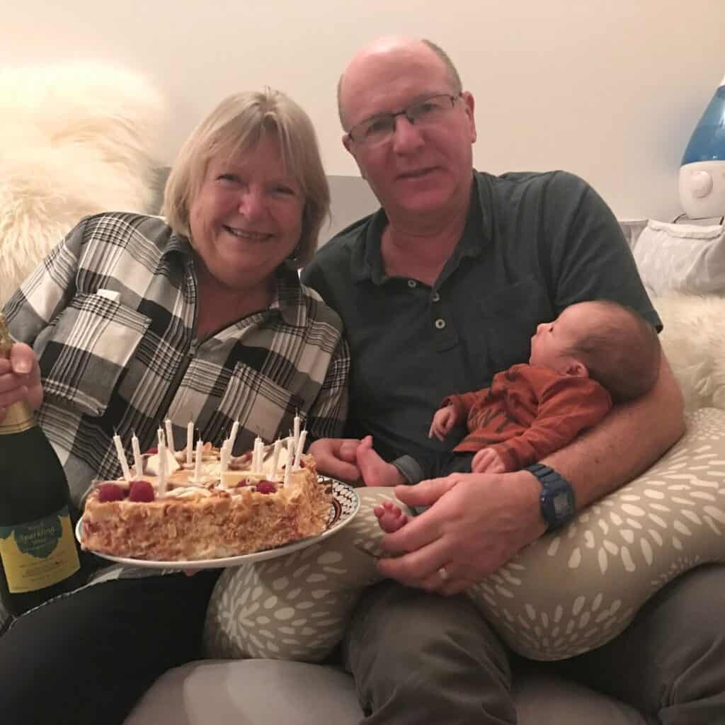Smiling grandparents seated on a cozy couch, holding a birthday cake with candles and a bottle of sparkling wine. The grandfather cradles a newborn baby in a patterned cushion, creating a warm, multigenerational celebration scene.