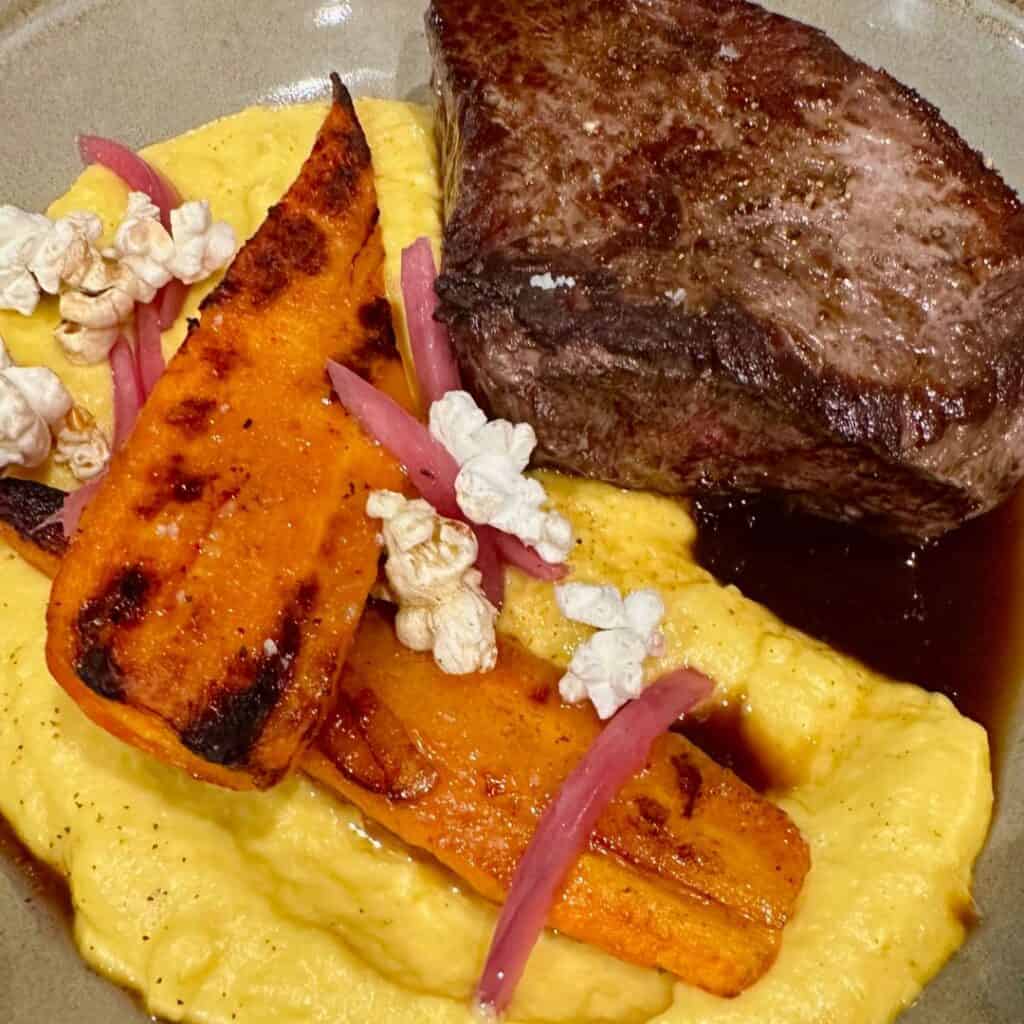Perfectly seared steak served with creamy polenta, charred carrot wedges, pickled red onions, and garnished with popcorn for a playful crunch. The plate blends rich, smoky, and tangy elements in a refined presentation.