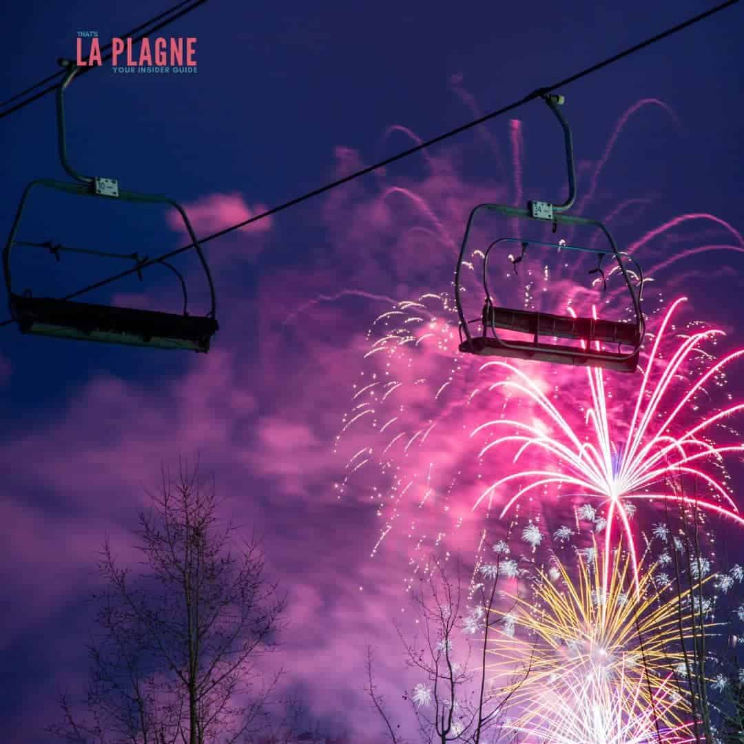 Ski lift chairs silhouetted against a dramatic night sky filled with pink, red, and gold fireworks at La Plagne. Bare tree branches frame the scene, adding contrast to the colorful burst of celebration.