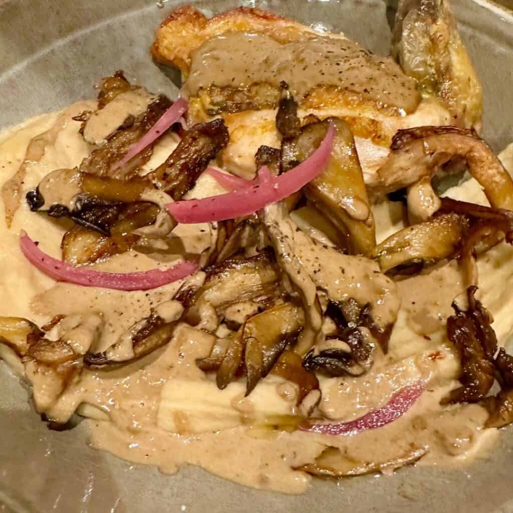 Pan-seared chicken breast served with a rich mushroom cream sauce, sautéed wild mushrooms, and pickled red onions. The dish is plated generously, showcasing earthy tones and a rustic, comforting presentation.