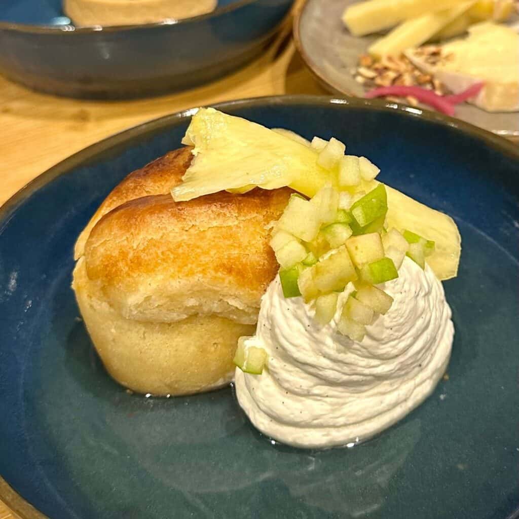 Golden, fluffy brioche served with a swirl of whipped cream, diced green apple, and slices of fresh pineapple. Presented in a deep blue dish with a light and airy plating style.
