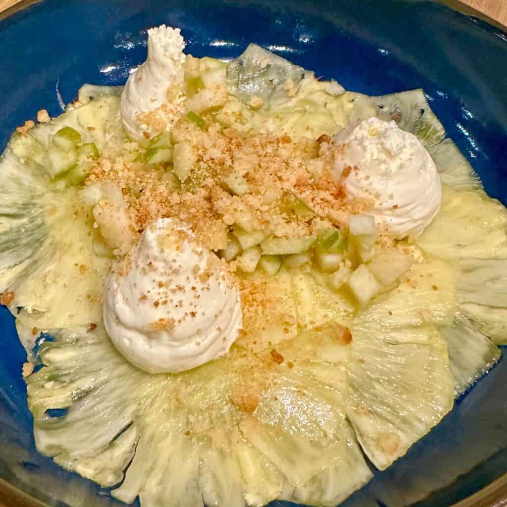 Paper-thin slices of pineapple arranged in a floral pattern, topped with whipped cream dollops, diced green apple, and a dusting of crumbled biscuit. A refreshing, tropical dessert with delicate textures.