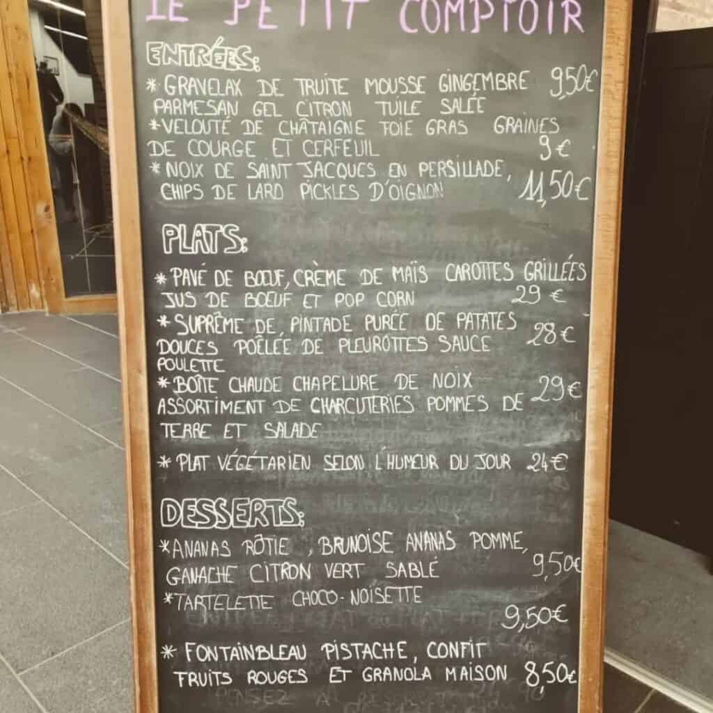 Chalkboard menu outside Le Petit Comptoir featuring a handwritten list of French entrées, main dishes, and desserts with prices. Highlights include dishes like velouté de châtaigne with foie gras, pavé de bœuf with corn cream, and roasted pineapple with lime ganache.