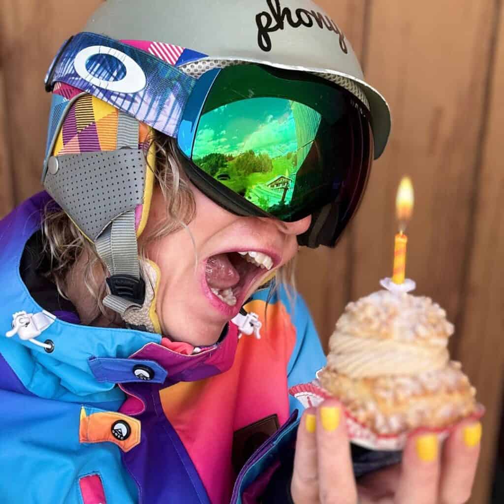 A person in a vibrant ski jacket and reflective goggles playfully pretends to take a big bite of a pastry topped with a lit birthday candle. Their ski helmet reads “phony,” and the reflection in their goggles shows a scenic mountain lodge and pine trees.