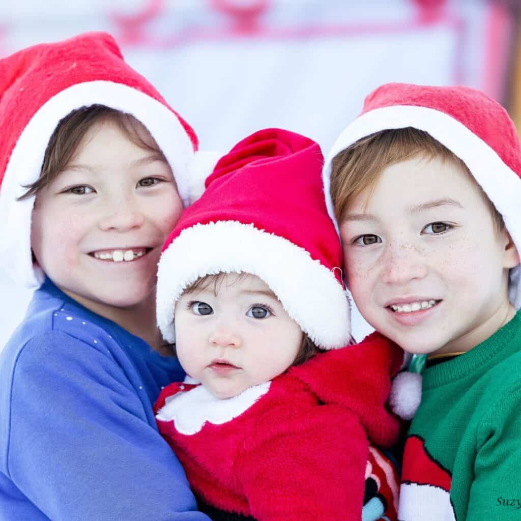Three young children dressed in cozy holiday outfits and red Santa hats pose together, smiling warmly. The older two gently hold their baby sibling, who wears a red fuzzy suit with a white collar, creating a joyful and festive Christmas portrait.