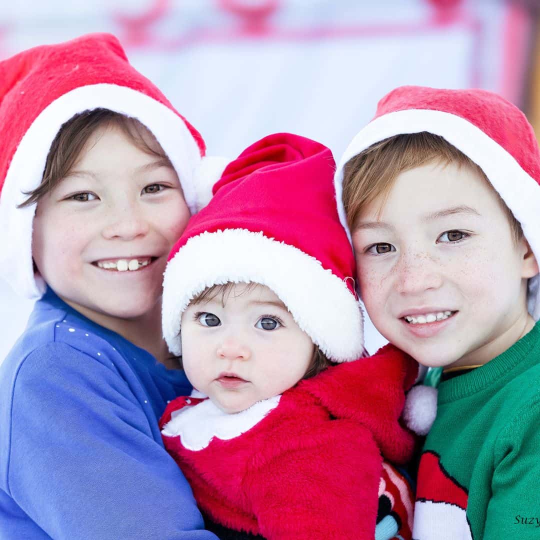 Three young children dressed in cozy holiday outfits and red Santa hats pose together, smiling warmly. The older two gently hold their baby sibling, who wears a red fuzzy suit with a white collar, creating a joyful and festive Christmas portrait.