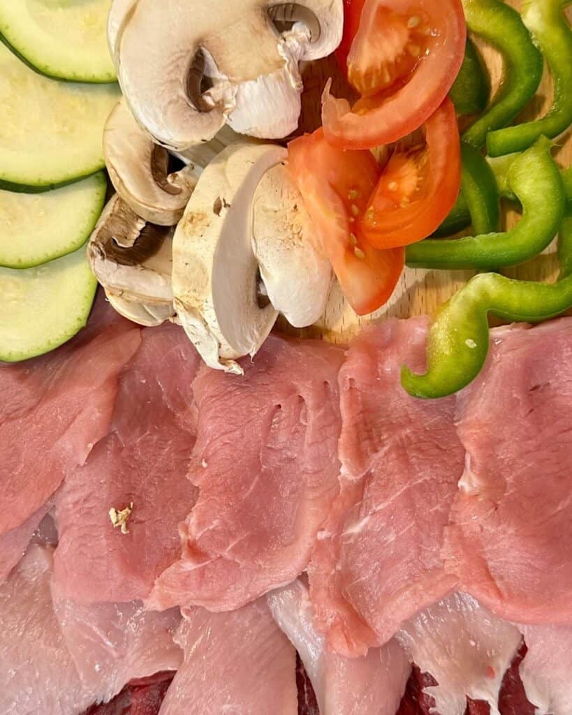 A colorful spread of fresh ingredients including thin slices of raw meat, zucchini, mushrooms, tomato wedges, and green bell pepper rings on a wooden cutting board.
