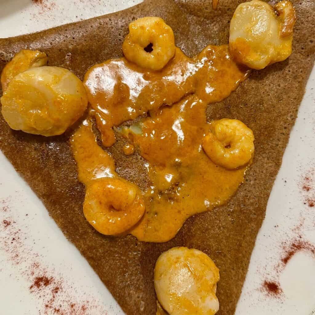 A savory buckwheat crepe folded into a triangle, topped with plump scallops and shrimp in a creamy orange sauce. The dish is served on a white plate with a light dusting of red spice around the edges for garnish.