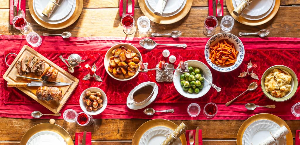 Top-down view of a beautifully set holiday dinner table with a red velvet runner and gold-accented place settings. The spread includes roast meats, Brussels sprouts, glazed carrots, roast potatoes, stuffing, gravy, and pigs in blankets.