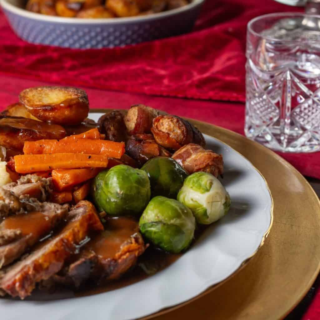 Close-up of a holiday dinner plate filled with roast pork and gravy, Brussels sprouts, glazed carrots, roast potatoes, and pigs in blankets. The plate sits on a gold charger with a red tablecloth and cut glass water tumbler beside it.