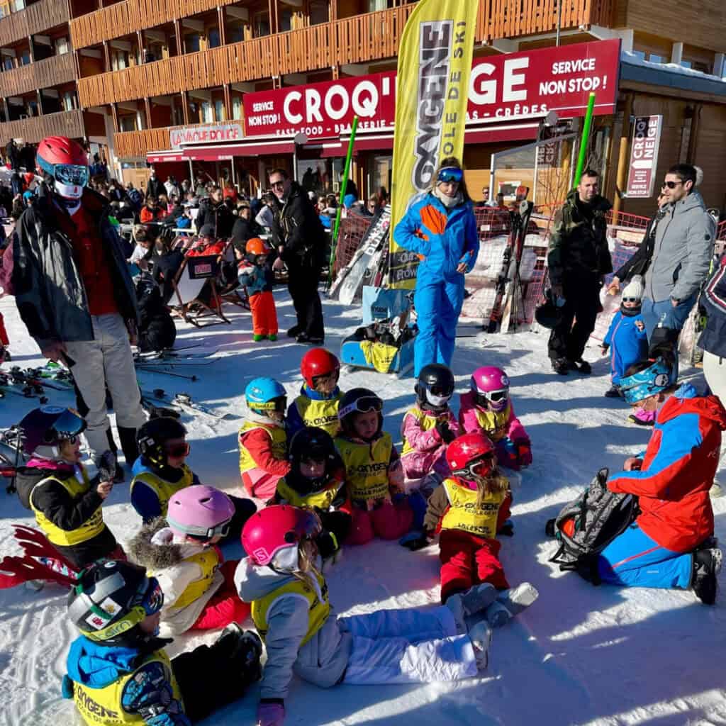 A lively ski resort scene with a group of young children in colorful ski gear and yellow Oxygène vests gathered on the snow for a lesson, surrounded by instructors and adults in front of a restaurant called "Croq’Neige."