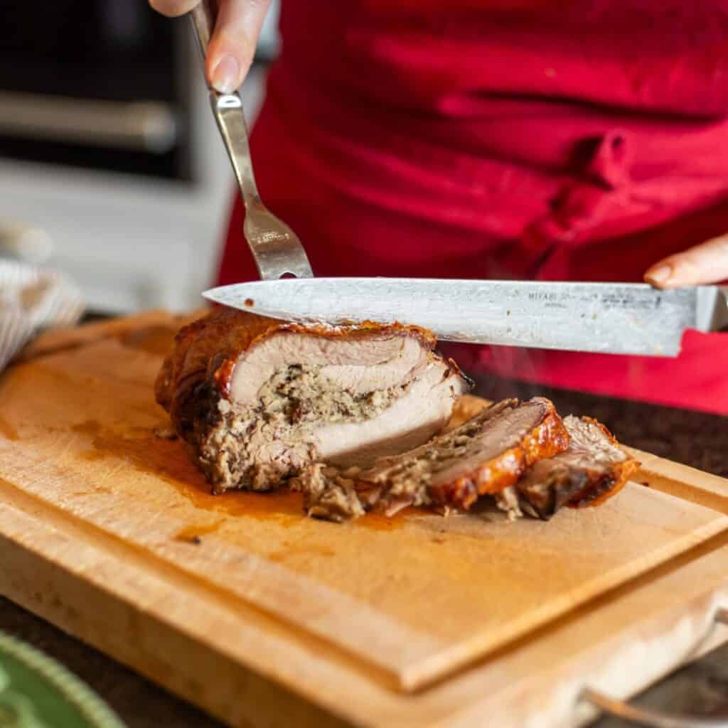A person in a red apron carves a juicy, herb-stuffed roast pork loin on a wooden cutting board. The meat is perfectly golden and tender, with savory juices visible on the board.