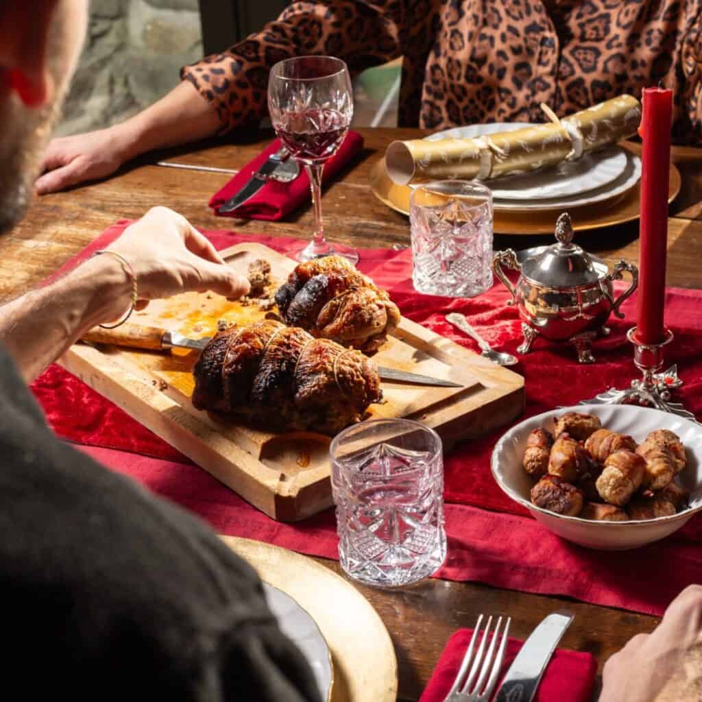 A cozy holiday table setting with a red tablecloth, crystal glasses, and a wooden cutting board holding a roast being sliced. Plates, crackers, and a bowl of pigs in blankets set the festive mood with warm lighting and rustic charm.