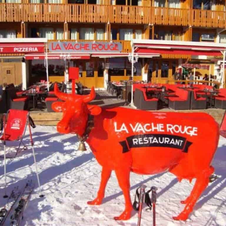 A large red cow statue marked “La Vache Rouge Restaurant” stands in front of a ski-in/ski-out restaurant with red-themed outdoor seating in La Plagne.