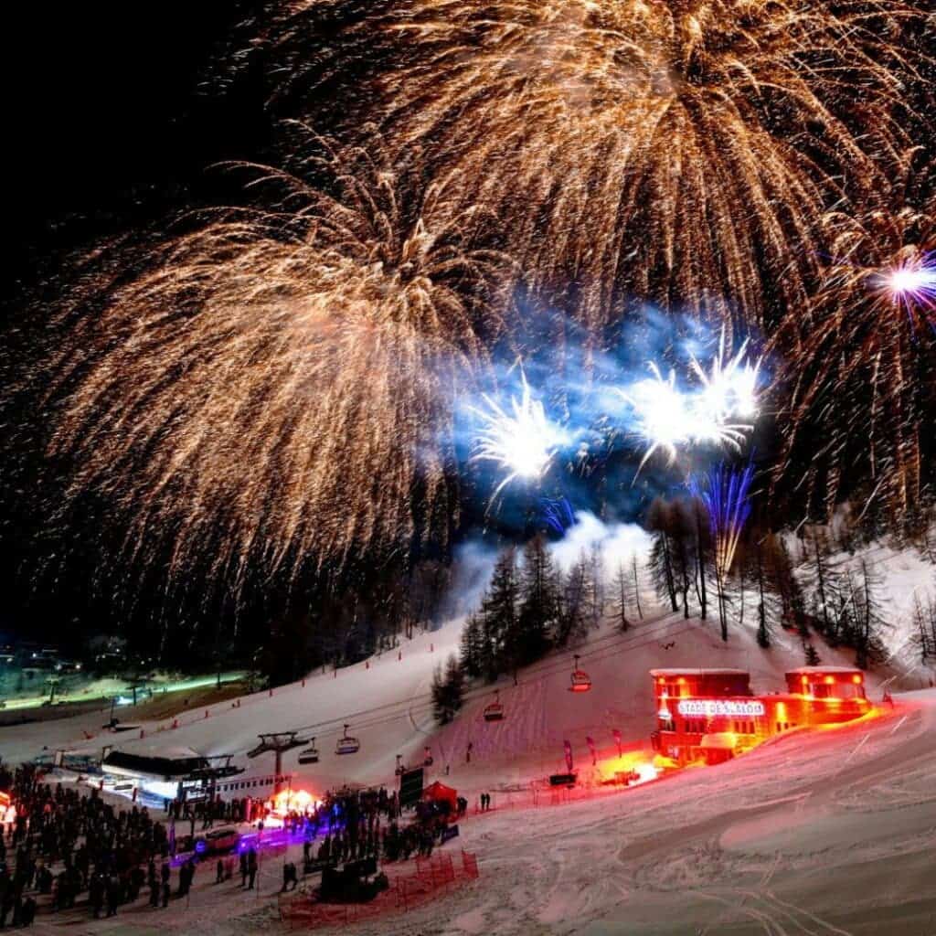 A vibrant night scene at a ski resort featuring a dazzling fireworks display over snowy slopes, with golden and blue bursts lighting up the sky. Crowds gather below near a brightly lit building labeled “TAPIS DES BLEUS,” surrounded by ski lifts and a forested hillside.