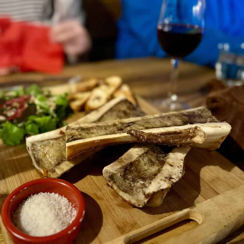 Roasted bone marrow served on a wooden board with a side of coarse salt, mixed greens, and baguette slices, with a glass of red wine in the background.