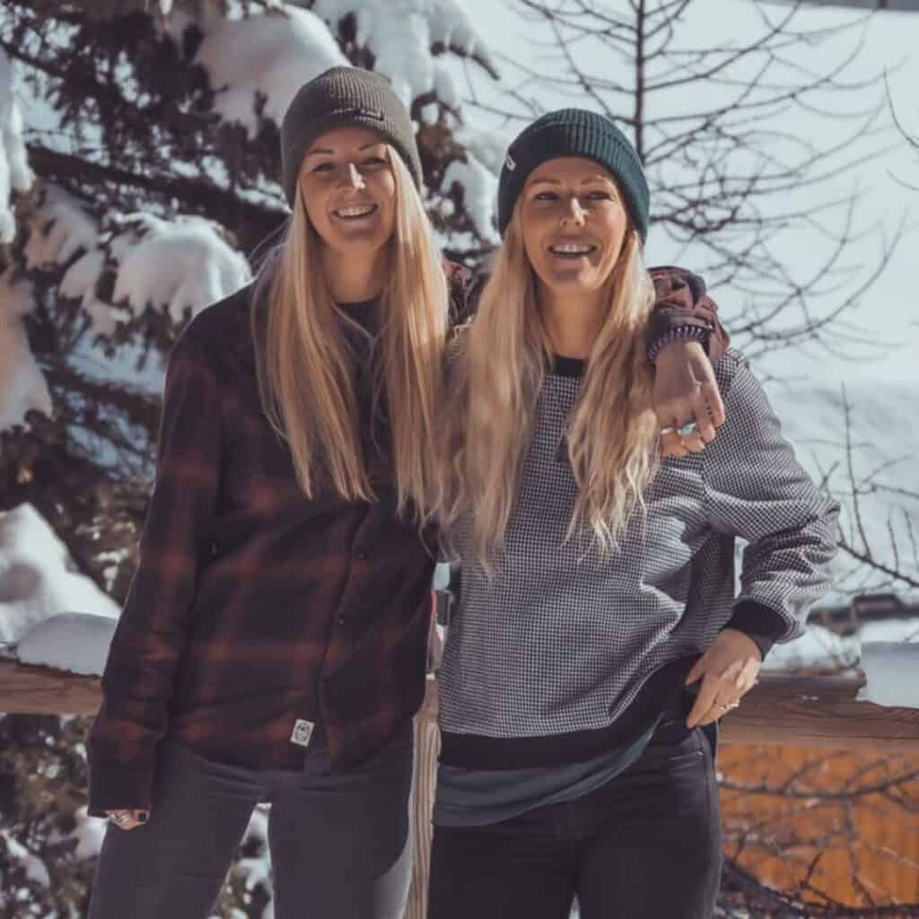 Two women dressed in cozy winter gear smile for the camera, standing arm-in-arm on a snowy balcony with frosted trees and soft mountain light in the background. Both wear beanies and casual winter layers.