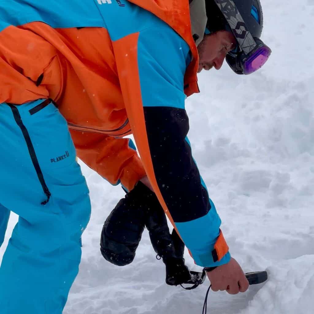 A ski patroller in a bright blue and orange uniform crouches in the snow while using an avalanche transceiver or beacon during a rescue or training exercise. Snow goggles rest on his helmet as he concentrates on the task.