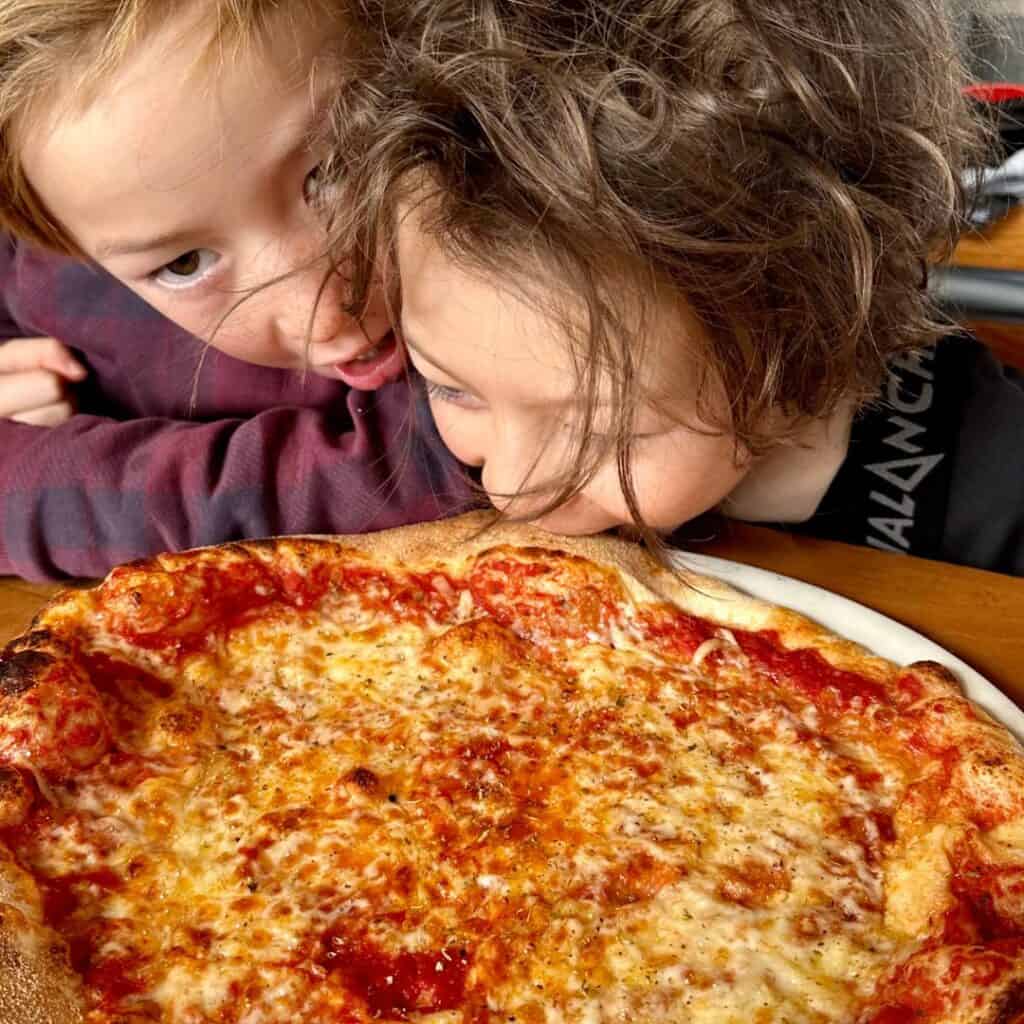 Two excited children lean eagerly over a large, cheesy pizza with a bubbly crust and rich tomato sauce, capturing a moment of joyful anticipation.