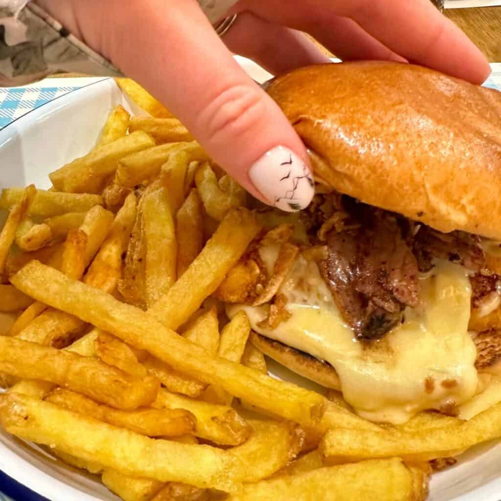 A hand holding a juicy burger with melted cheese and grilled meat, served in a bowl alongside golden, crispy French fries.
