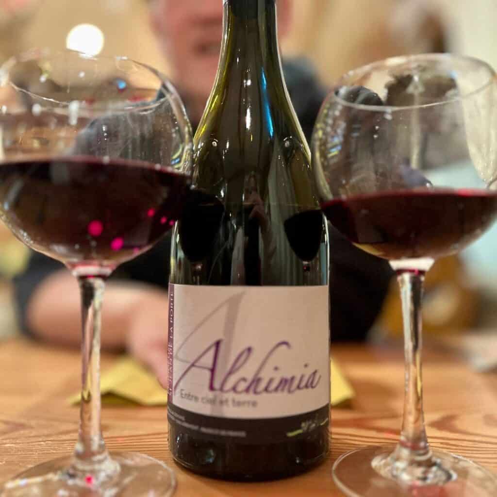 A bottle of red wine labeled "Alchimia" placed between two glasses of red wine on a wooden table, ready for a cozy dinner setting.
