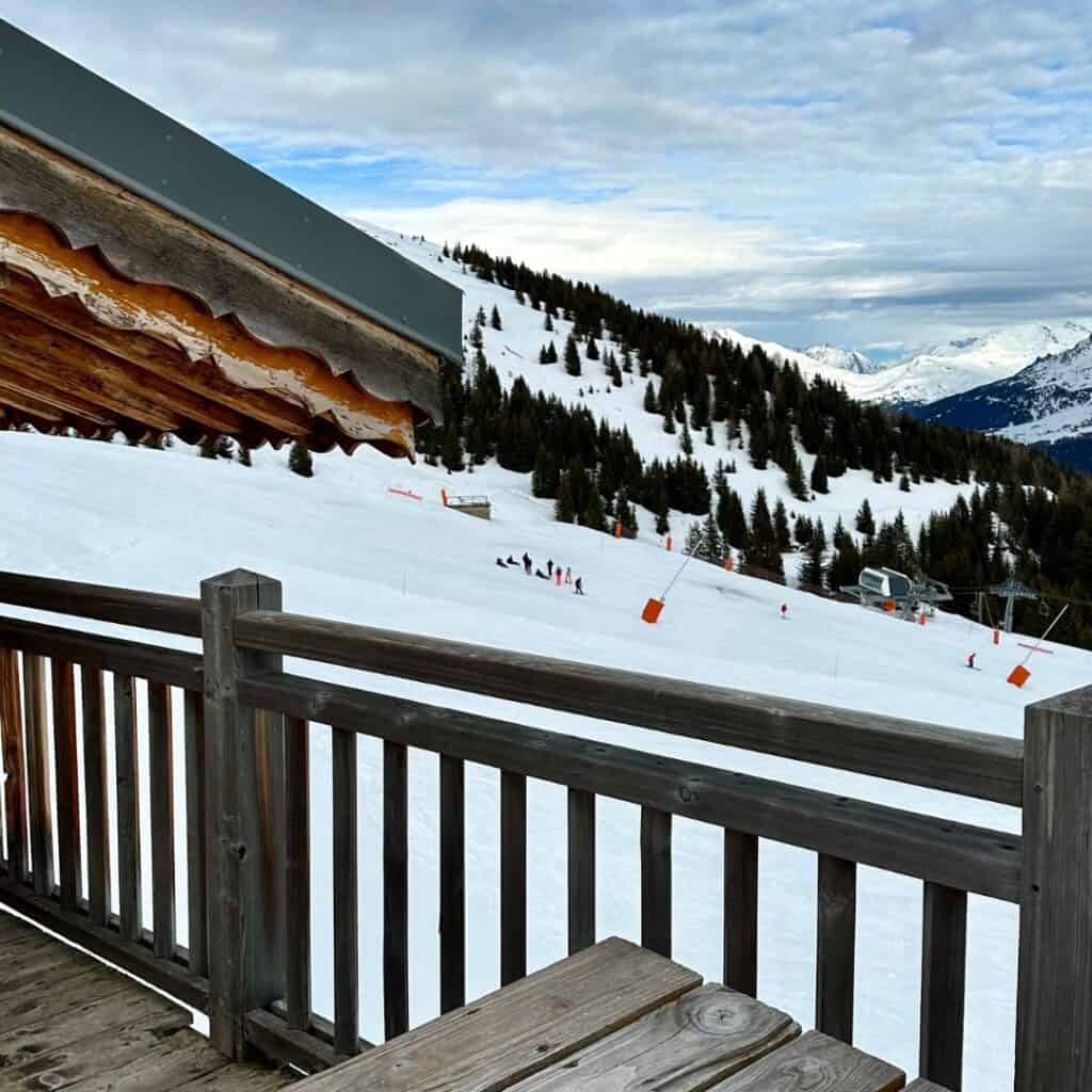View from a wooden deck looking out onto a snowy ski slope lined with pine trees, with a small group of skiers in the distance. A ski lift and orange slope markers are visible, set against a backdrop of snow-covered mountains under a cloudy sky. The edge of a wooden alpine lodge roof frames the top left corner.