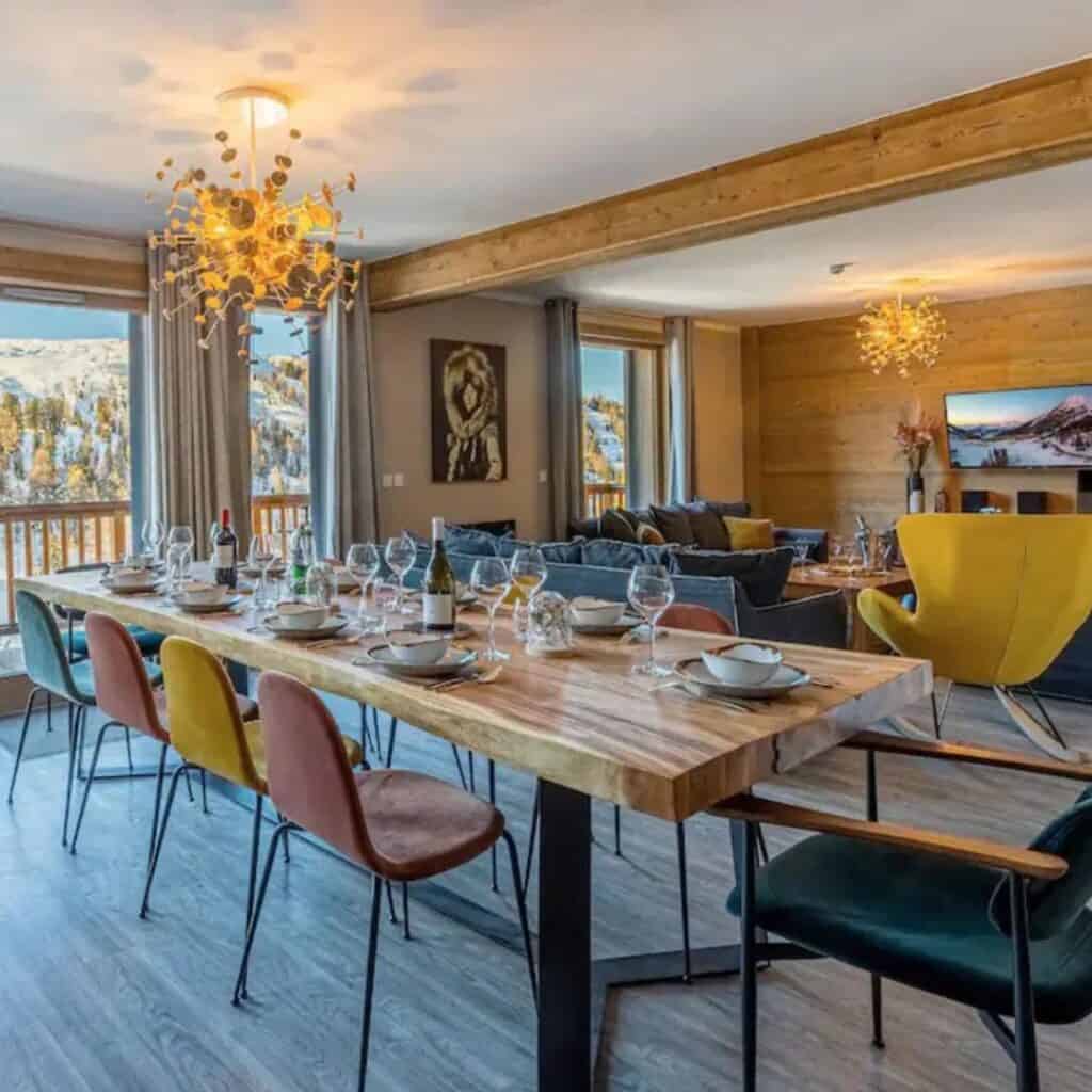 Spacious open-plan dining room with a long wooden table set for a meal, surrounded by colorful modern chairs. Large windows offer views of the snow-covered mountains, and the space is lit by chic pendant lights.