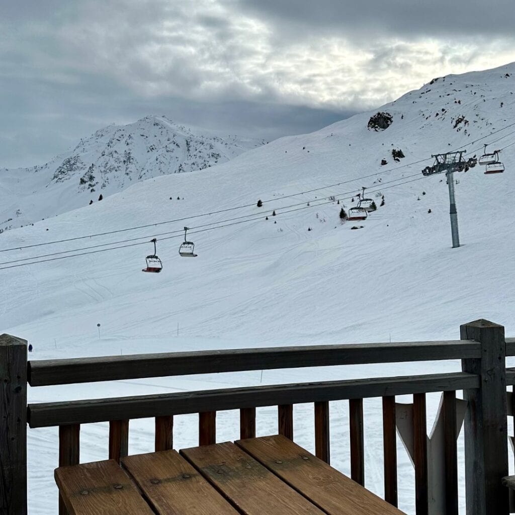 Scenic view from a wooden deck at Le Carroley in La Plagne, looking out at snow-covered slopes with chairlifts suspended in motion under a moody, overcast sky. A wooden picnic table sits in the foreground, framing the snowy mountain vista.