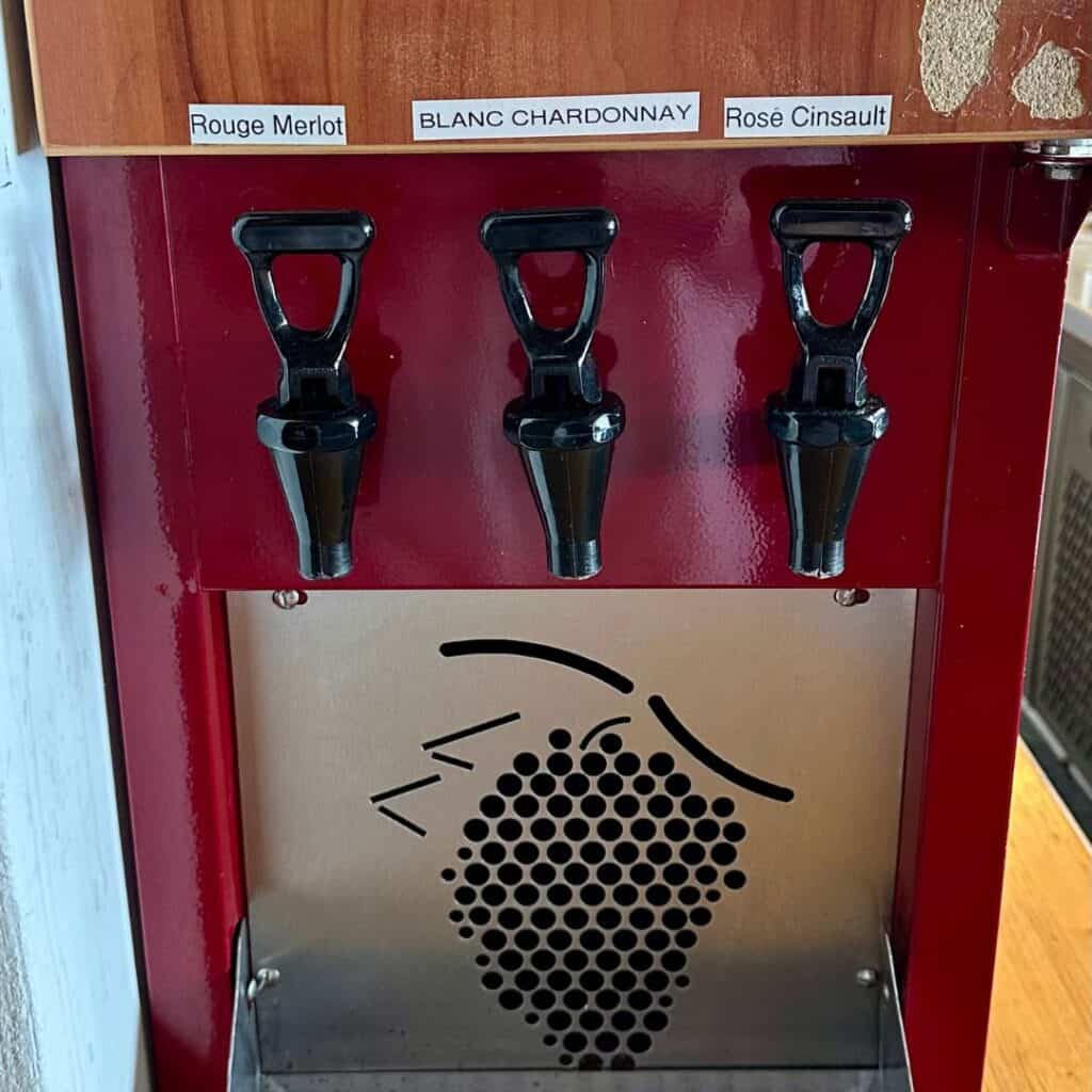 A wine dispenser machine with three taps labeled Rouge Merlot, Blanc Chardonnay, and Rosé Cinsault, set against a shiny red panel with a grape cluster graphic below.