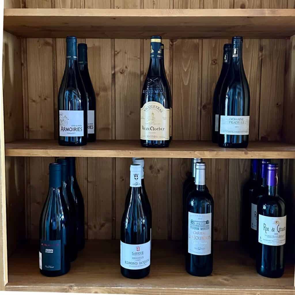 A wooden shelf stocked with various bottles of red wine, including labels such as Armoiries, Veux Clocher, Domaine Pradelle, and Savoie Edmond Jacquin. The rustic wood backdrop adds to the cozy alpine aesthetic.