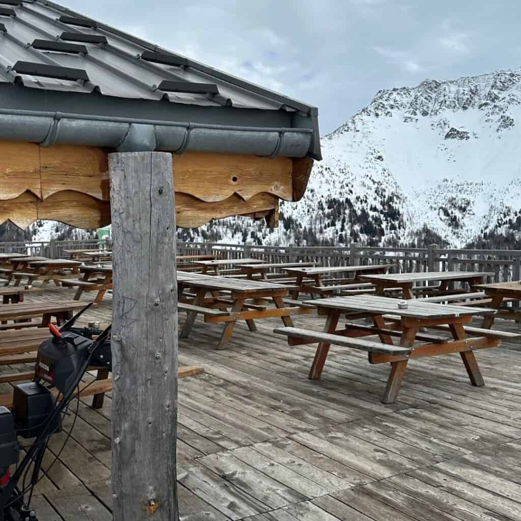 Wooden terrace filled with empty picnic tables at Le Carroley in La Plagne, overlooking snowy alpine slopes and a rugged mountain backdrop. A wooden post and snowblower are visible in the foreground beneath the chalet roof.
