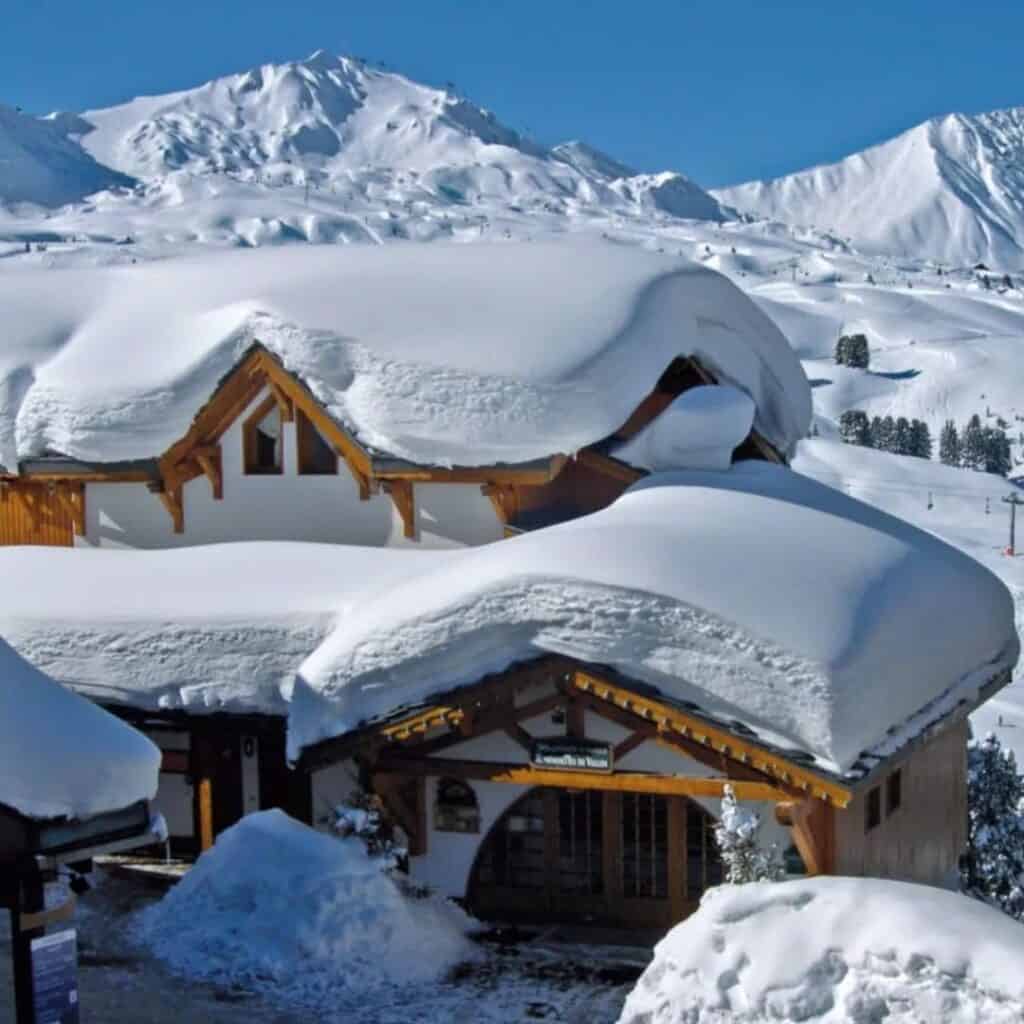 Charming wooden chalets blanketed in thick layers of snow with a backdrop of sunlit alpine mountains. The heavy snowpack creates a cozy, postcard-perfect winter village feel.