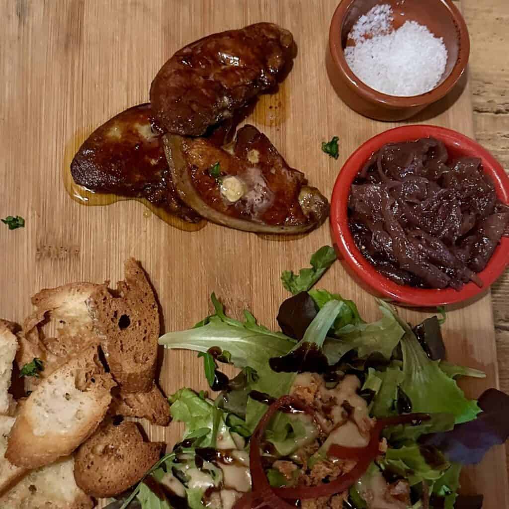 A rustic wooden board topped with seared foie gras, caramelized onions, a sprinkle of sea salt, mixed greens, and crispy toasted baguette slices.